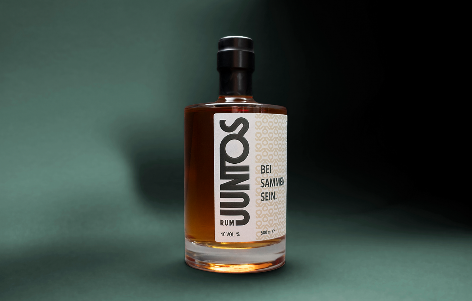 JUNTOS Rum Produktfoto. Eine Flasche frontal mit seitlicher Drehung, fotografiert, damit man das Logo auf dem Etikett gut sehen kann, in einem dunkelgrünen Setting.